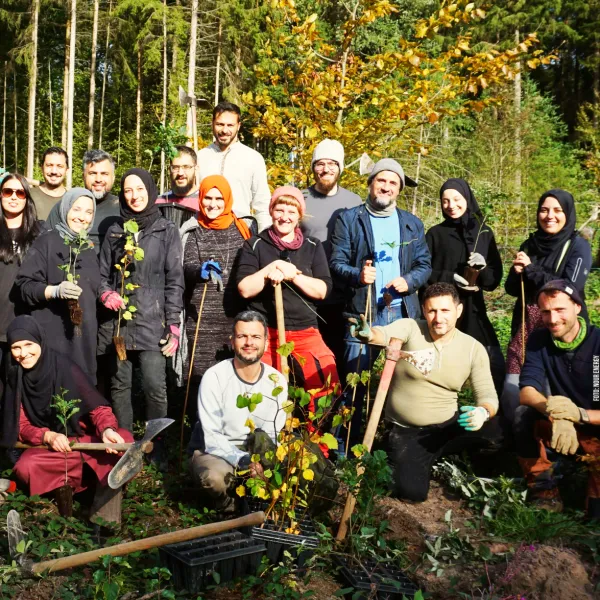 Gruppe von Menschen im Wald mit Setzlingen und Werkzeug, manche tragen Kopftuch oder Mütze